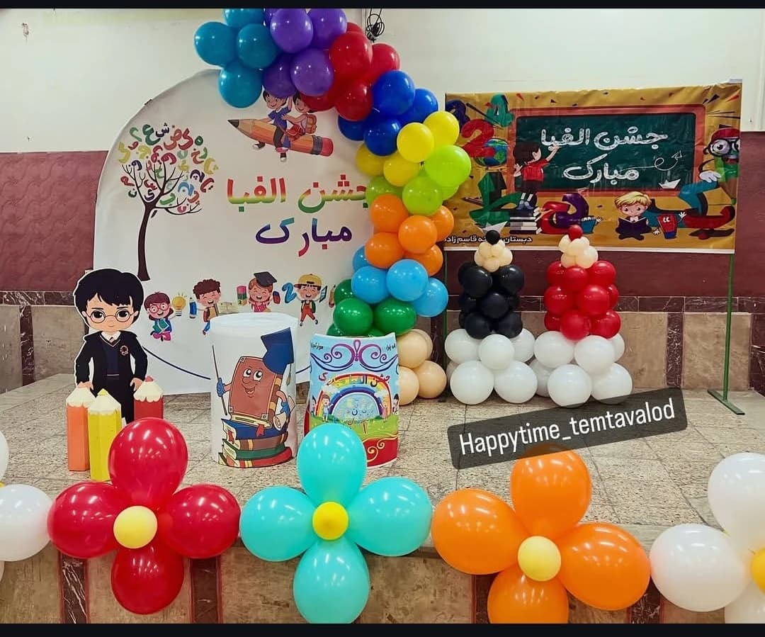 دکوراسیون جشن الفبا با طاق بادکنکی رنگینکمانی، پنل درخت حروف الفبا، کتاب خندان، گلهای بادکنکی و ستونهای قلاب روی پسزمینه چوبی