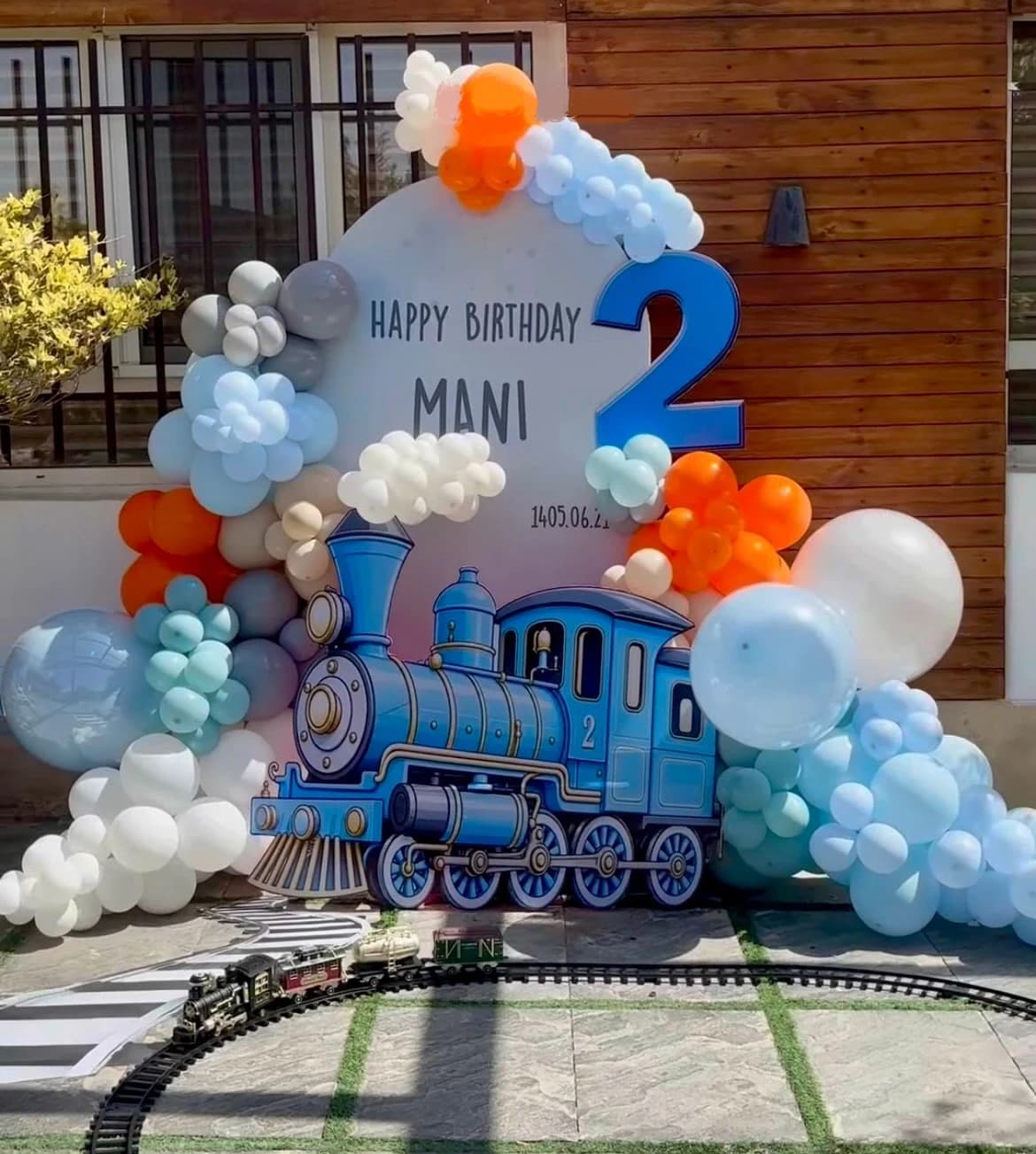دکور تولد ۲ سالگی مانی با تم قطار شامل بکدراپ دایرهای سفید با نوشته Happy Birthday و نام Mani، عدد آبی ۲، استند لوکوموتیو آبی کارتونی، آرک بادکنکی آبی و نارنجی و سفید و قطار اسباببازی روی ریل در فضای باز